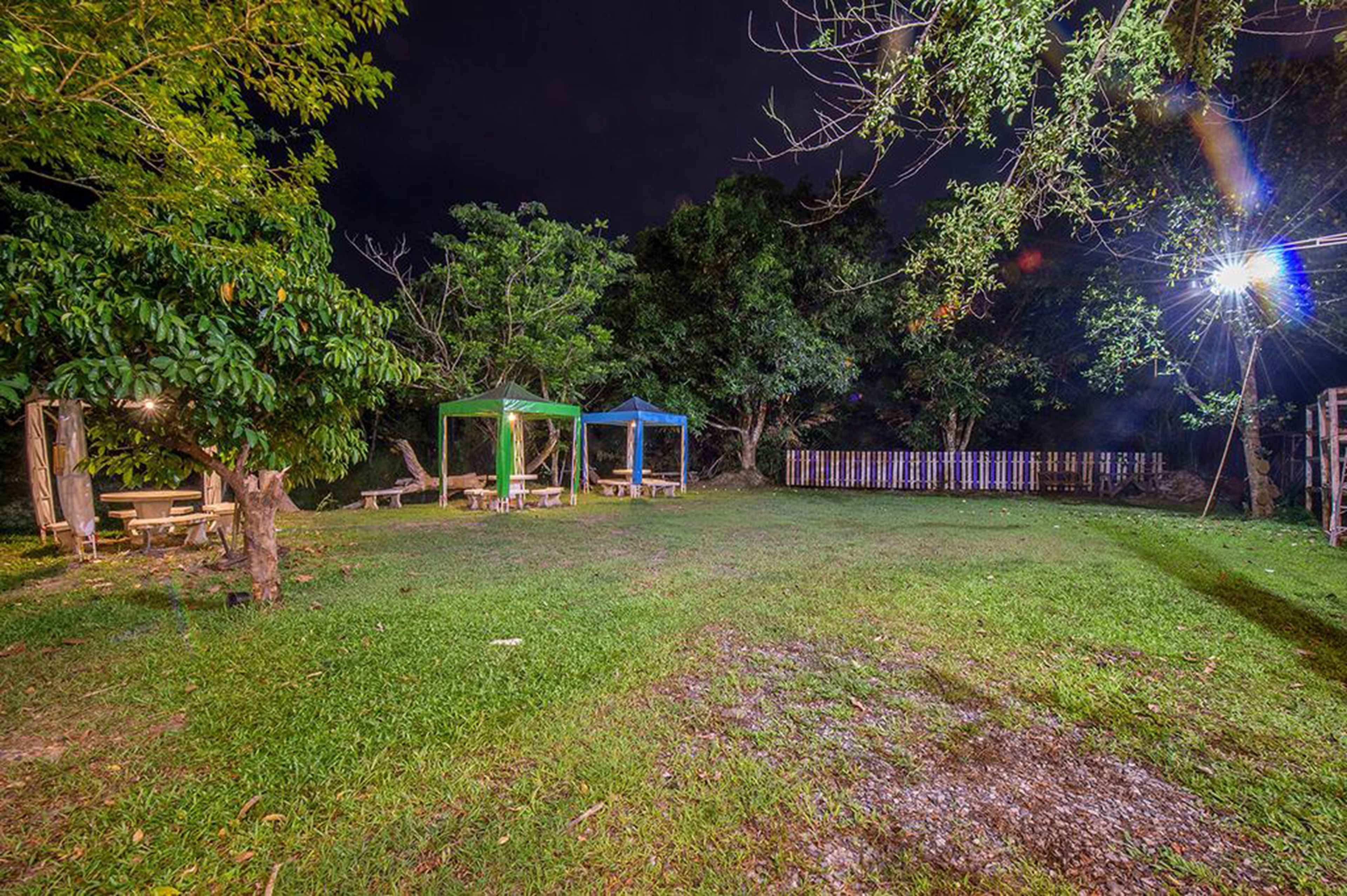 Un espacio exterior bien iluminado con mesas de picnic, una zona de estar cubierta y una cerca de madera.