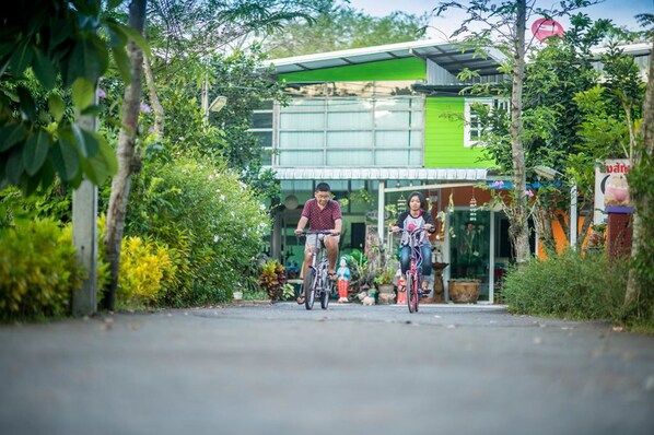 A street scene with two people riding bicycles. There is a building with a green facade and large windows in the background. - Poomvarin Resort (Nakhon Nayok)