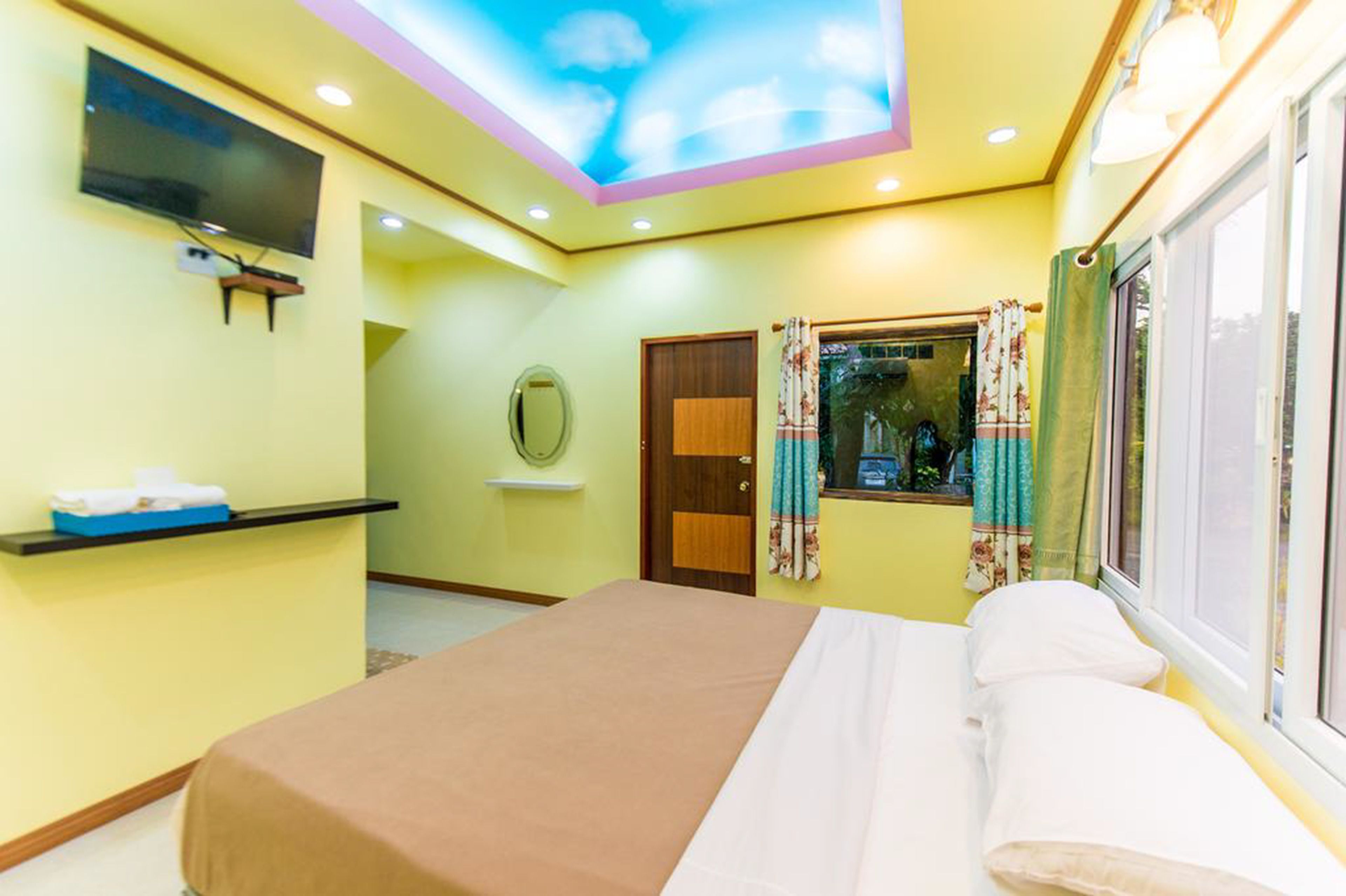Une chambre d’hôtel avec un lit, un téléviseur fixé au mur, une fenêtre avec des rideaux et un plafond décoré de motifs de nuages.