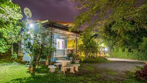 A small, rustic outdoor seating area with a table and benches, surrounded by lush greenery and a pathway leading to a building with a satellite dish. - Poomvarin Resort (Nakhon Nayok)