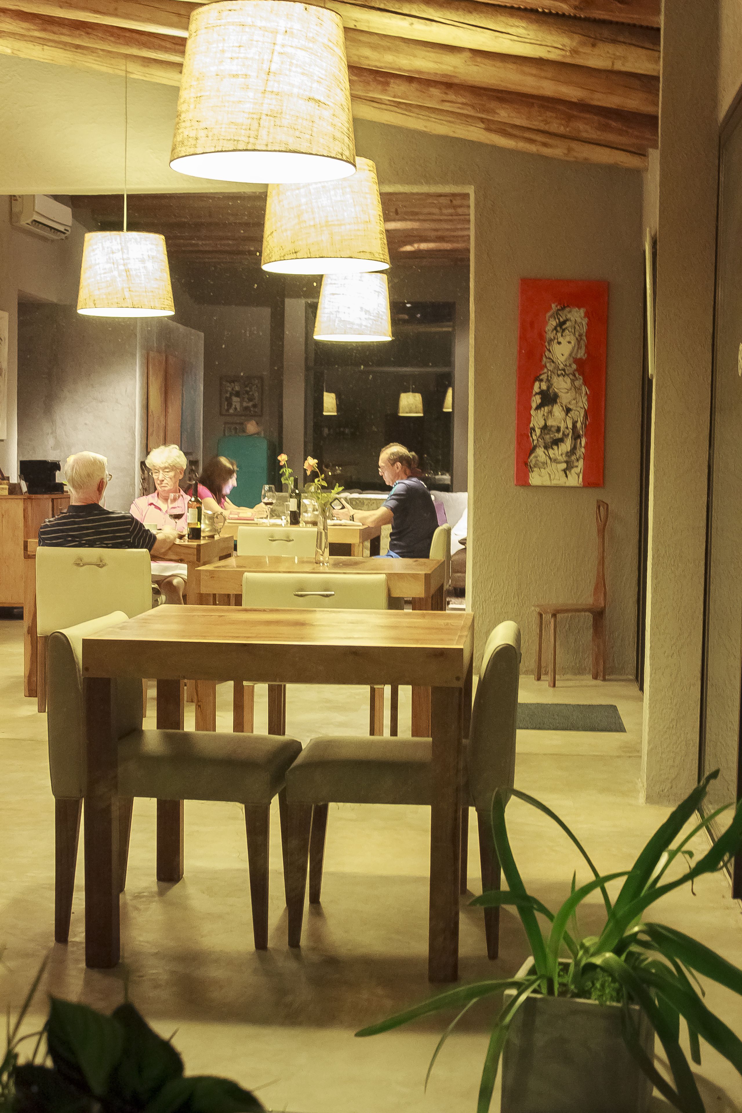 A dining area with wooden tables and chairs, hanging lamps, and a red artwork on the wall.