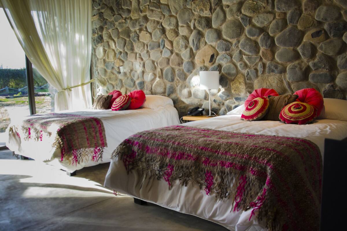 Deux lits avec du linge de lit blanc et des couvertures roses et brunes, un mur en pierre et une lampe.
