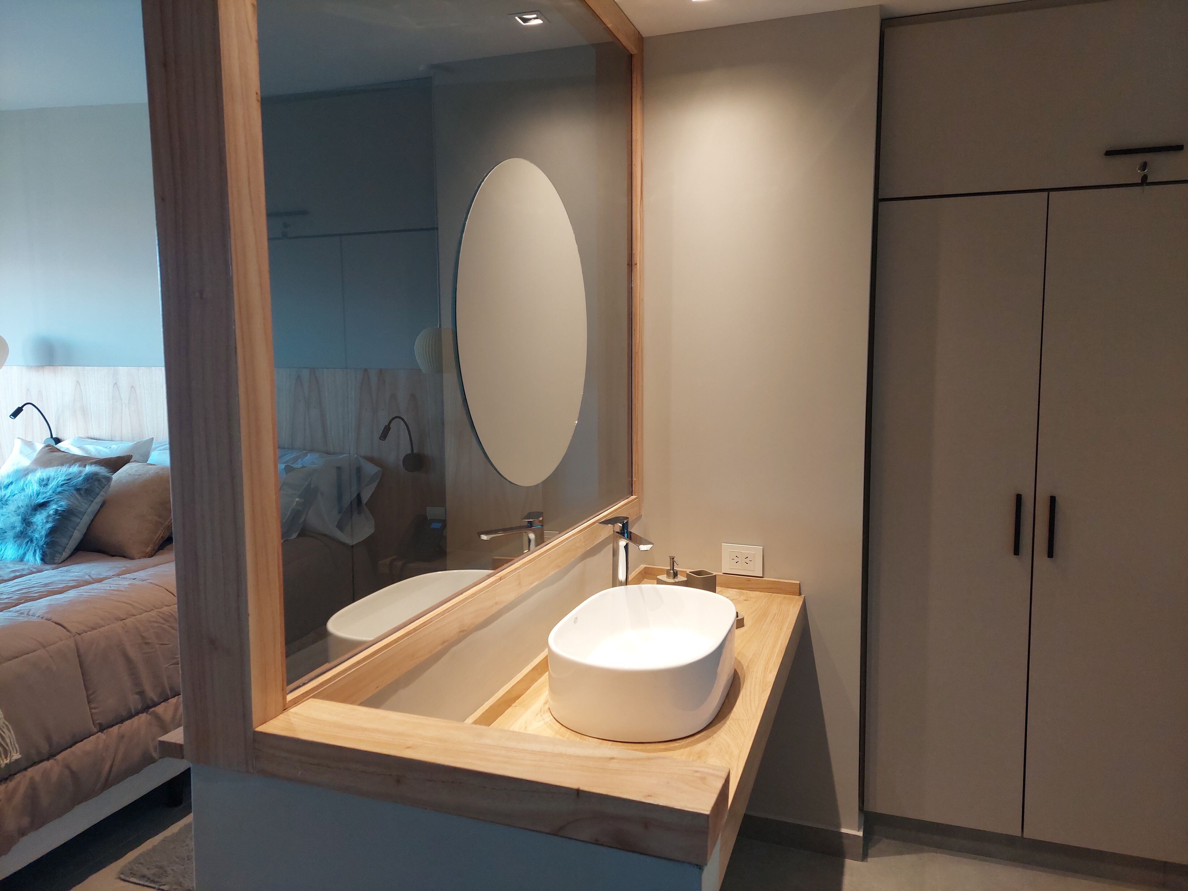 A modern bathroom with a wooden vanity, a large mirror, and a bed with a headboard in the background.