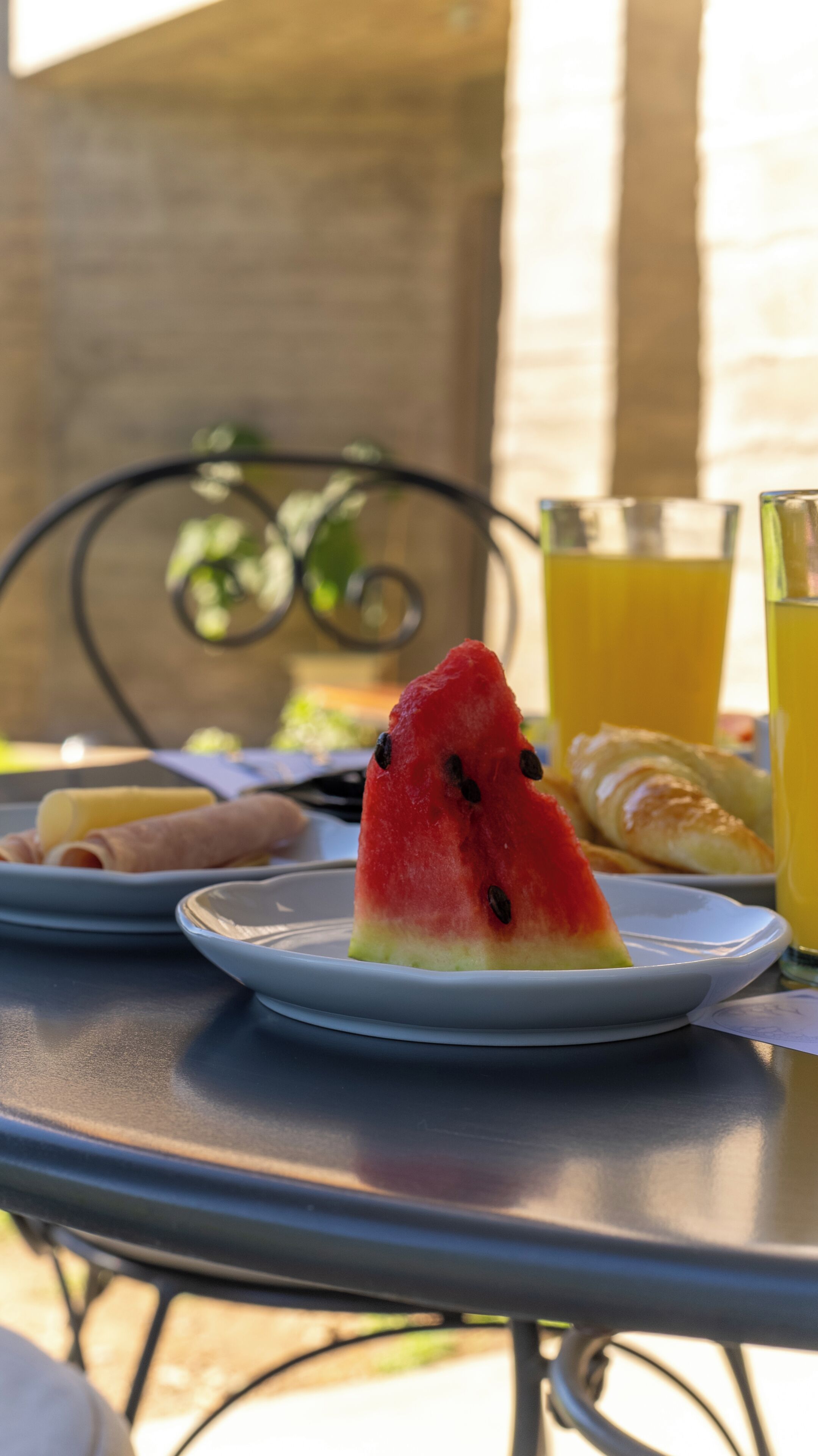 A slice of watermelon on a plate, a croissant, and a glass of orange juice on an outdoor table.