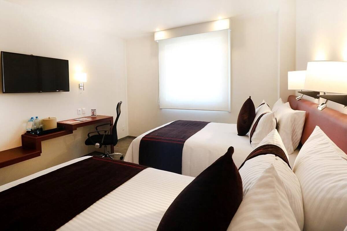 Une chambre d’hôtel avec deux lits, un bureau, une chaise, une télévision et une fenêtre avec des stores.