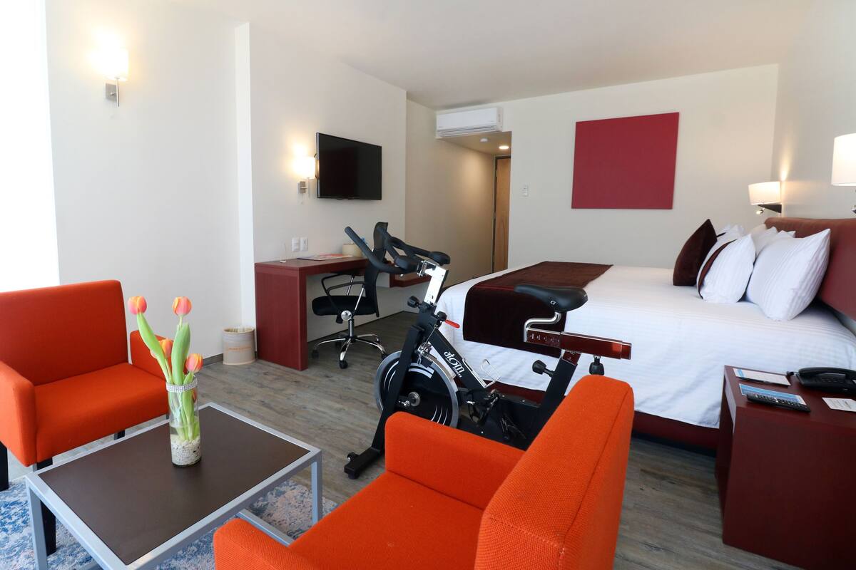 Une chambre d’hôtel avec un lit, deux fauteuils orange, un bureau, une télévision et un vélo d’appartement.
