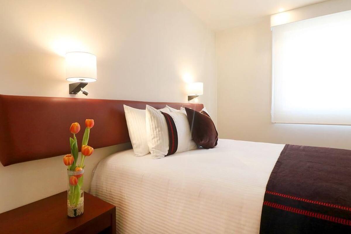 Une chambre d’hôtel avec un grand lit, deux lampes de chevet, une table de chevet ornée d’un vase de tulipes et une fenêtre avec des stores.