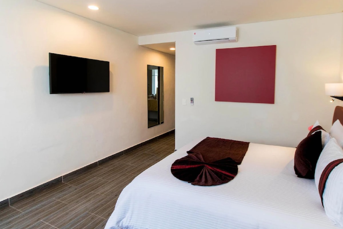 A hotel room with a bed, a flat-screen TV mounted on the wall, a red accent wall, and a hallway leading to another room.