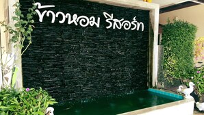 Pared negra con escritura tailandesa blanca, una pequeña piscina y plantas decorativas.