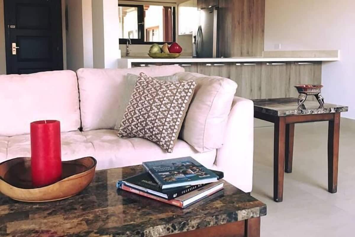 A living room with a white sofa, a coffee table with books and a red candle, and a kitchen in the background.