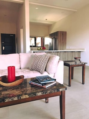 A living room with a white sofa, a coffee table with books and a red candle, and a kitchen in the background. - Hotel Gran Balcón (Granada)