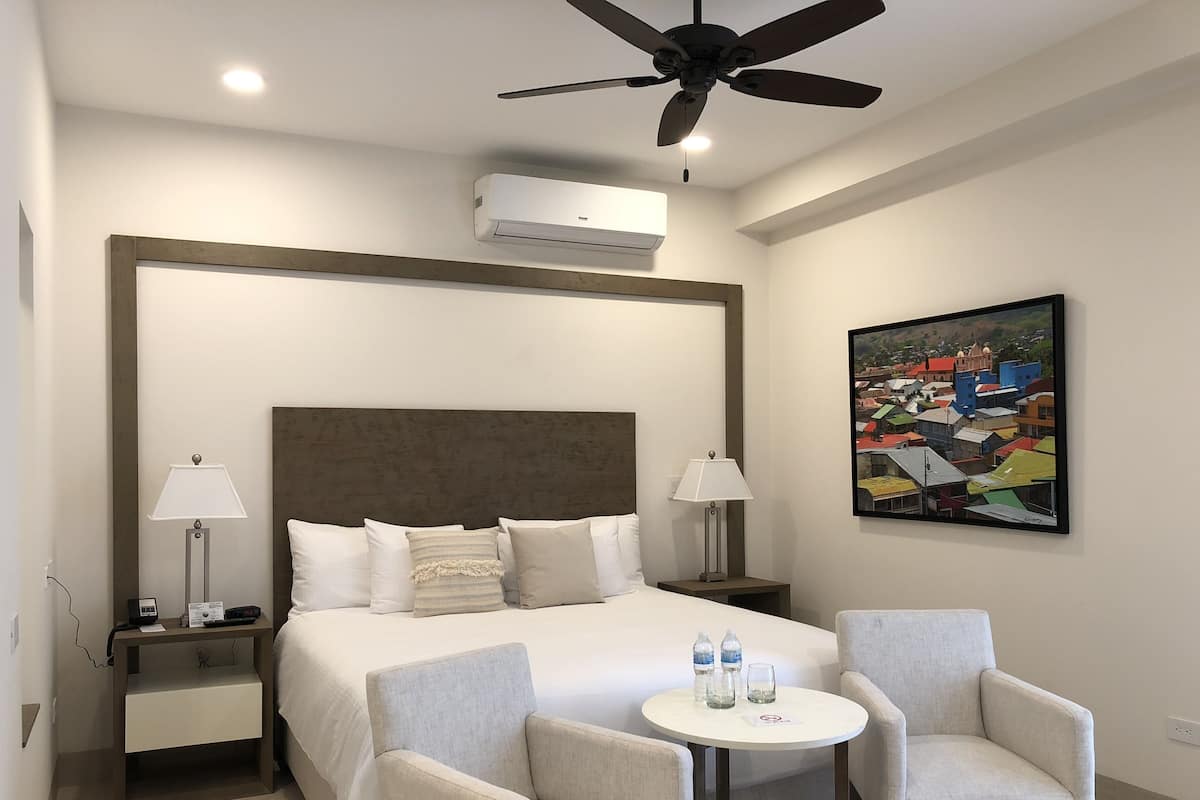 A hotel room with a large bed, two bedside tables with lamps, a ceiling fan, an air conditioning unit, a small round table with two chairs, and a colorful painting on the wall.