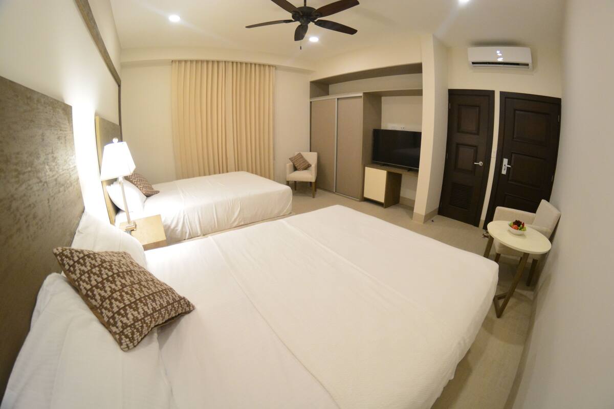 A hotel room with two beds, a TV, a small table with a bowl of fruit, a chair, and an air conditioning unit.