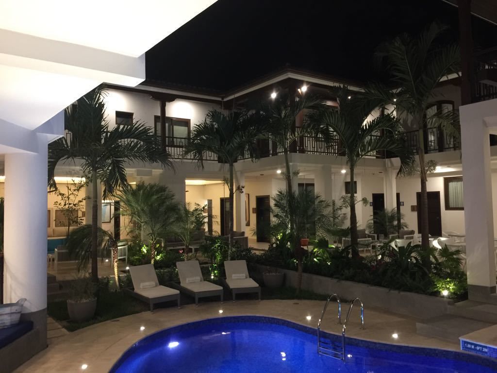 Zona de piscina con sillones de descanso y palmeras, rodeada por un edificio de varios pisos con balcones.