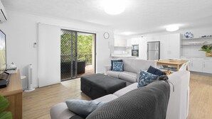 A modern living room with a sofa, ottoman, and a dining table. There is a kitchen area with cabinets and a refrigerator. A sliding glass door leads to an outdoor area with a patio table and chairs. - Aria Del Mare (Coolangatta)