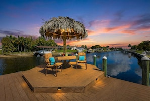 A wooden deck with a thatched umbrella, two chairs, a table, and a view of a water body with houses in the background.