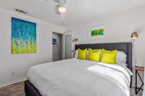 Ein modernes Schlafzimmer mit einem großen Bett, leuchtend gelben Kissen, einem grauen Kopfteil und Bildern an den Wänden.