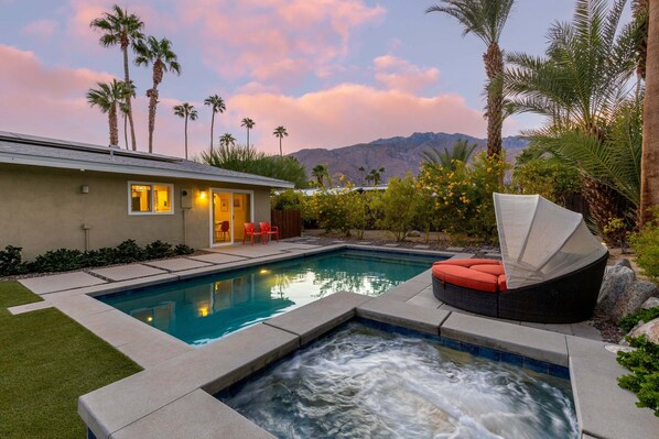 A swimming pool with a hot tub, a house with a patio, and palm trees.