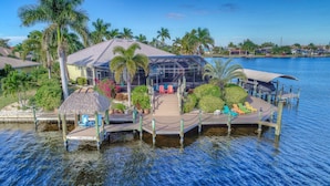 A waterfront property with a glass-enclosed structure, a dock with lounge chairs, and a thatched-roof pavilion. - Waterfront Paradise | Pool, Spa, Kayaks & Dock (Cape Coral)