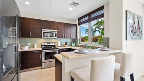 A modern kitchen with dark cabinets, stainless steel appliances, a central island, and a window with a view of palm trees. - Hemingway 3 Bedroom Condo 2 Blocks from the Beach (Deerfield Beach)