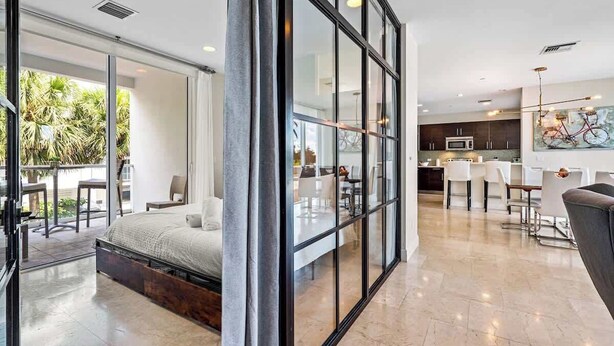 A modern interior with a glass partition, a bed, a dining area, and a kitchen. - Hemingway 3 Bedroom Condo 2 Blocks from the Beach (Deerfield Beach)