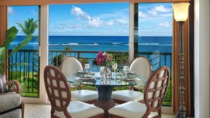 A dining table with four chairs, a vase with flowers, and a view of the ocean through large windows.