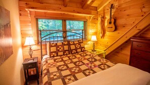A cozy bedroom with a wooden ceiling, a bed with a quilt, two bedside lamps, a guitar on the wall, and a window with blinds. - Rock n Rhapsody - Blue Ridge Log Cabin - Minutes to West Jefferson - Fireplace - Foosball (West Jefferson)