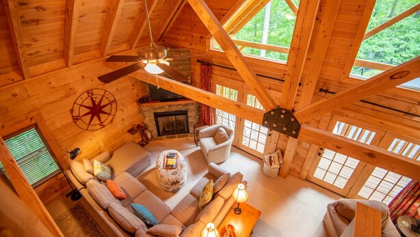 A cozy log cabin interior with a fireplace, a ceiling fan, and a large window overlooking greenery. - Rock n Rhapsody - Blue Ridge Log Cabin - Minutes to West Jefferson - Fireplace - Foosball (West Jefferson)