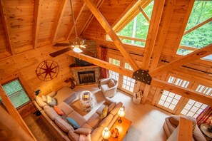 A cozy log cabin interior with a fireplace, a ceiling fan, and a large window overlooking greenery.