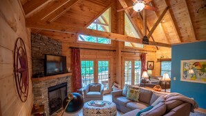 A cozy log cabin interior with a stone fireplace, a television mounted above it, and a large window overlooking a deck. - Rock n Rhapsody - Blue Ridge Log Cabin - Minutes to West Jefferson - Fireplace - Foosball (West Jefferson)