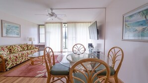 Een woonkamer met een bank, een eettafel met glazen blad en stoelen. Er is een plafondventilator, een aan de muur bevestigde televisie en een raam met gordijnen.