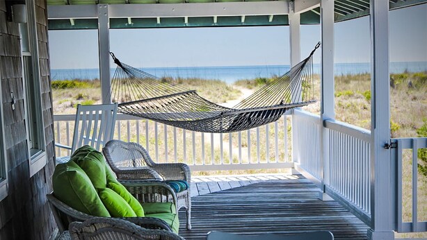 A covered porch with wicker chairs and a hammock overlooking a beach.