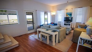 A spacious living room with a wooden floor, a sofa set, a television, and a balcony with a view of the beach.