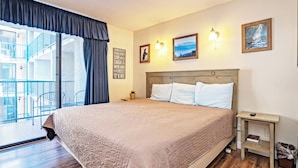 Habitación de hotel con una cama grande, una mesita de noche pequeña, una lámpara de pared y vistas al balcón exterior.