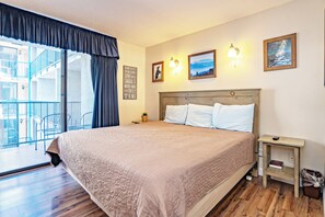 Habitación de hotel con una cama grande, una mesita de noche pequeña, una lámpara de pared y vistas al balcón exterior.