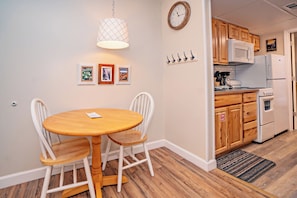 Una cocina con armarios de madera, un refrigerador blanco y un microondas. Hay una mesa de comedor con dos sillas y una lámpara colgante.