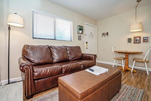 Ein Wohnzimmer mit braunem Ledersofa, passendem Hocker, einer Lampe, einem Esstisch mit Stühlen und einem Fenster mit Jalousien.
