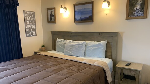 A neatly made bed with a brown quilt, a bedside table with a remote control, and wall-mounted light fixtures.