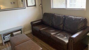 A living room with a brown leather sofa, a small wooden side table, and a bench. There is a floor lamp and a window with blinds.