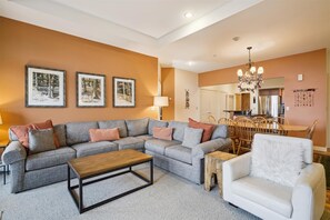 A living room with a sectional sofa, a coffee table, a white armchair, and a dining area with wooden chairs and a chandelier.