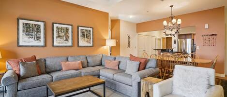 A living room with a sectional sofa, a coffee table, a white armchair, and a dining area with wooden chairs and a chandelier.