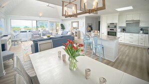 A modern kitchen with white cabinets, a central island, and a dining area with a white table and chairs. - Wrightsville Beach Coastal Retreat (Wrightsville Beach)