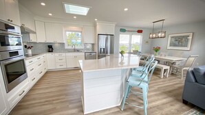 A modern kitchen with white cabinetry, a central island, and a dining area with light blue chairs. - Wrightsville Beach Coastal Retreat (Wrightsville Beach)