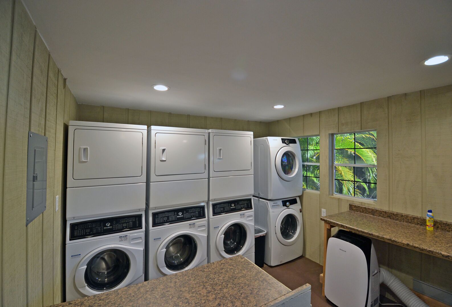 A laundry room with stacked washers and dryers, a granite countertop, and a window with a view of greenery.