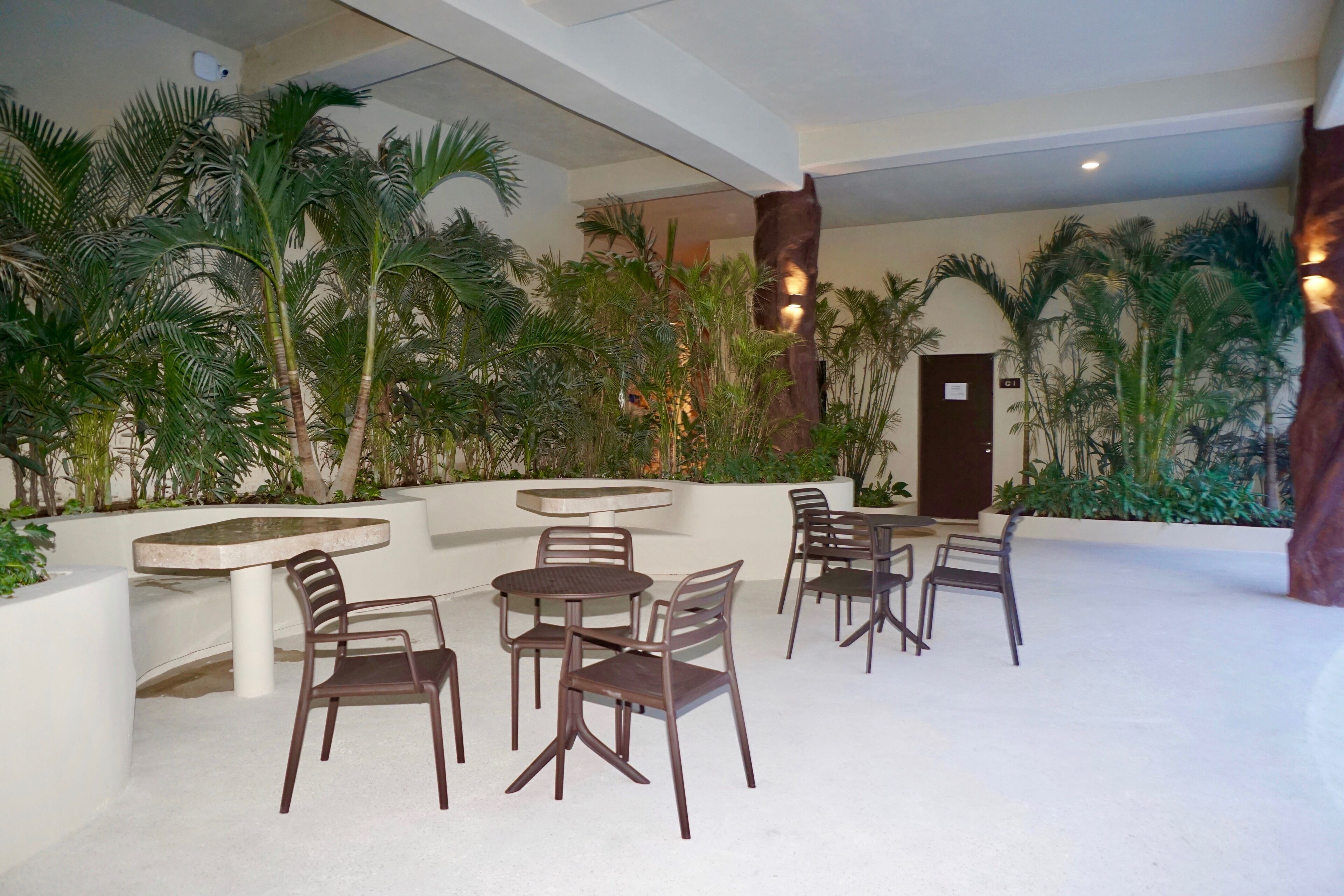 A spacious indoor area with multiple seating arrangements, including tables and chairs, surrounded by lush greenery and palm trees.