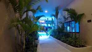 A corridor with a concrete floor, surrounded by potted plants and palm trees, leading to a building with a balcony. - 1 Bedroom * HighSpeedConnection* Downtown quite & safe-5th ave steps away (Playa del Carmen)