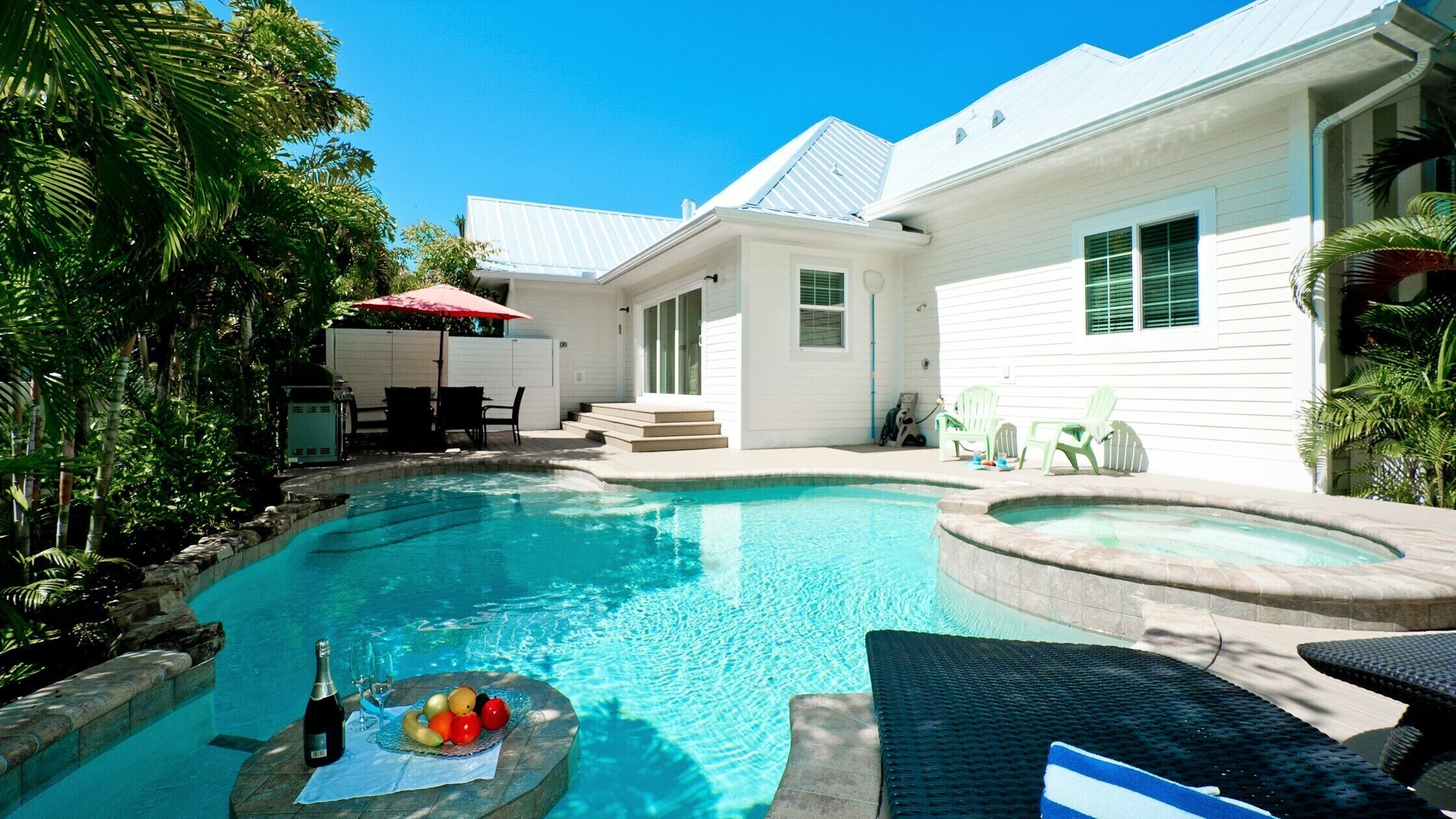 Un jardin avec une piscine, une terrasse dotée de meubles d’extérieur, et une maison aux murs recouverts de bardage blanc et aux volets verts.