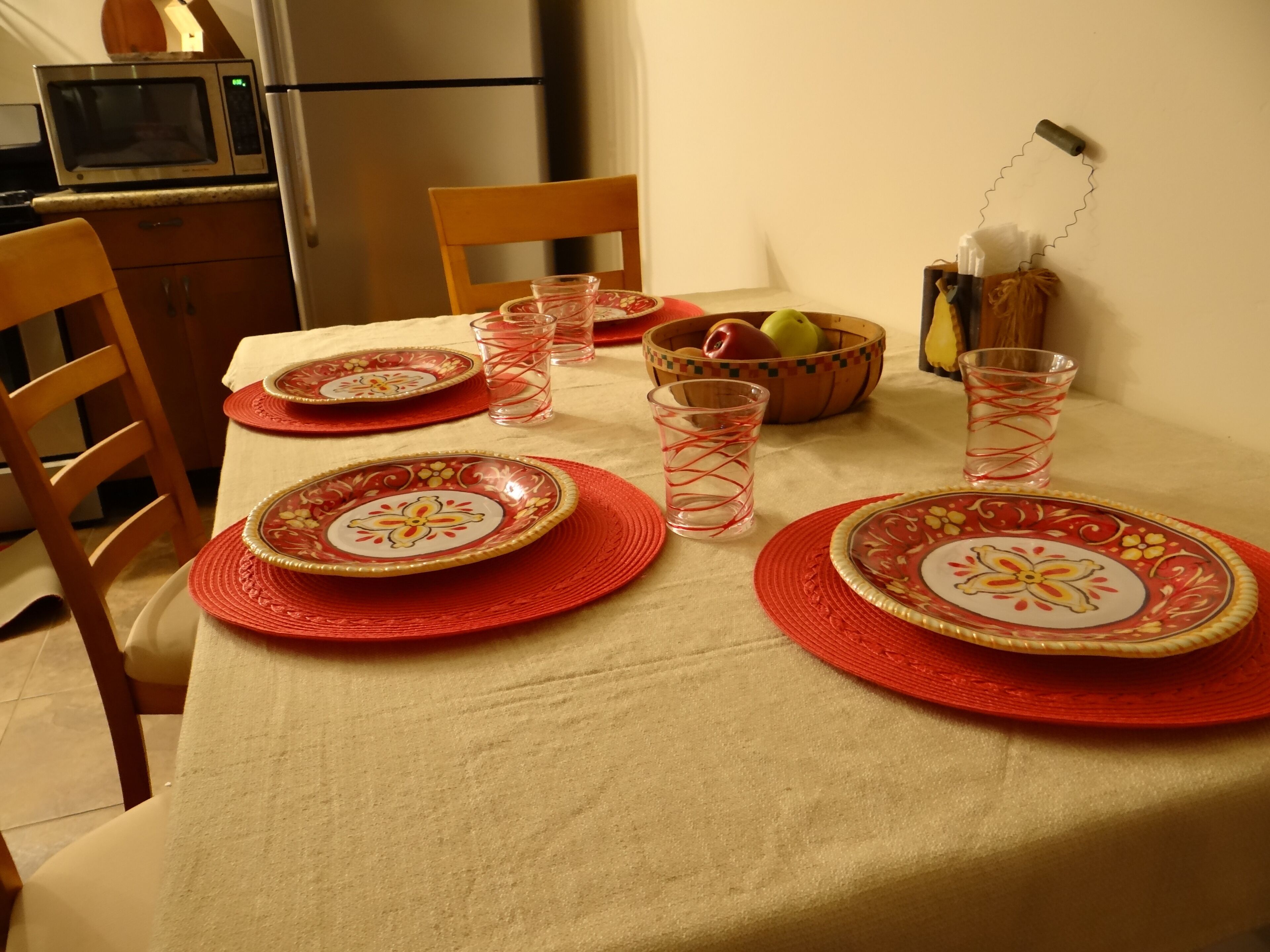 Une table à manger est dressée avec des sets de table rouges, des assiettes et des verres transparents. Un saladier rempli de fruits et un rouleau d’essuie-tout sont posés sur la table.