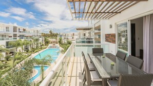 Un balcón con mesa y sillas de vidrio con vista a una piscina y edificios cercanos.