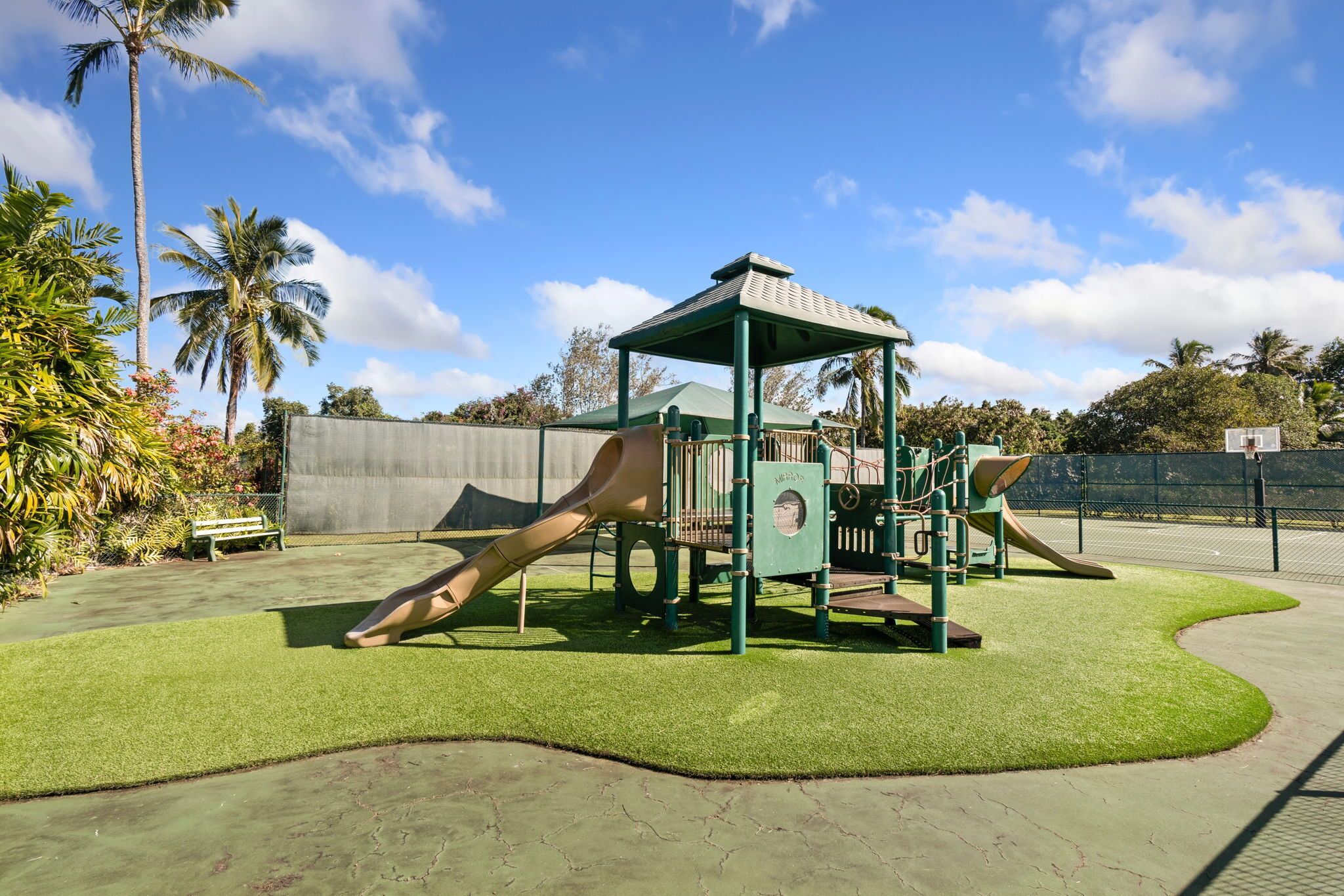 A playground with a slide and climbing structures, surrounded by a fence and palm trees.